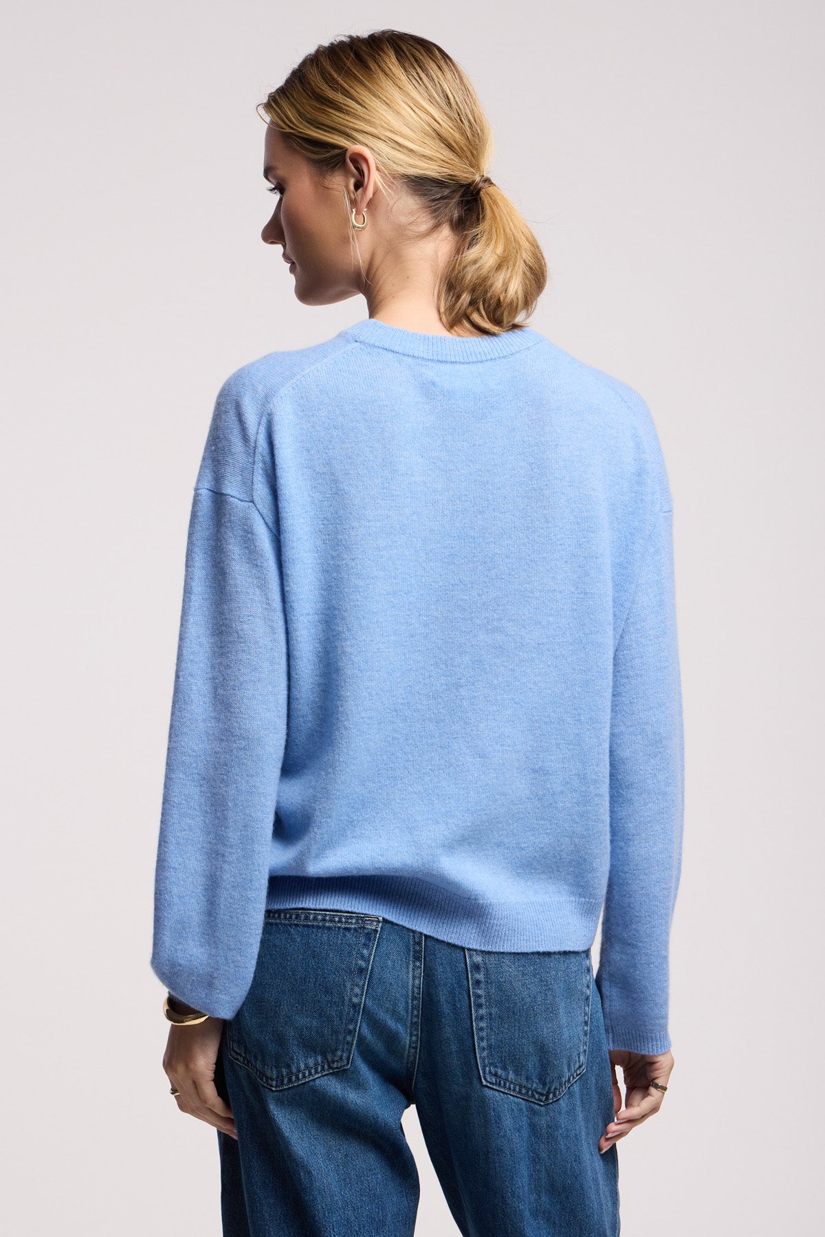 A person stands with their back to the camera, wearing the Velvet by Graham & Spencer BRYNNE CASHMERE SWEATER in light blue with ribbed trims, paired with blue jeans.-40521782493377