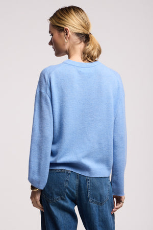 A person stands with their back to the camera, wearing the Velvet by Graham & Spencer BRYNNE CASHMERE SWEATER in light blue with ribbed trims, paired with blue jeans.
