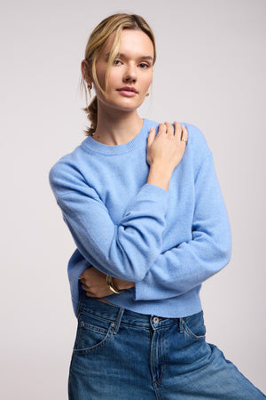 A woman wears the BRYNNE CASHMERE SWEATER by Velvet by Graham & Spencer in light blue with ribbed trims and blue jeans, standing against a plain background with one arm crossed and the other hand on her shoulder.