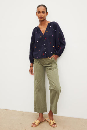 A woman standing in a studio wearing a navy blouse with colorful polka dots, Velvet by Graham & Spencer Mya Cotton Canvas Pants, and tan sandals.