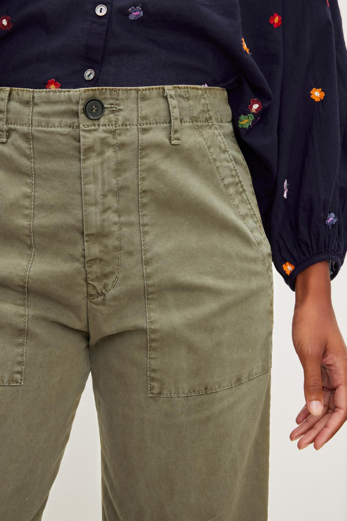   Close-up of a person wearing Velvet by Graham & Spencer's MYA COTTON CANVAS PANT and a black shirt with floral embroidery, focusing on the pants and a hand by the side. 