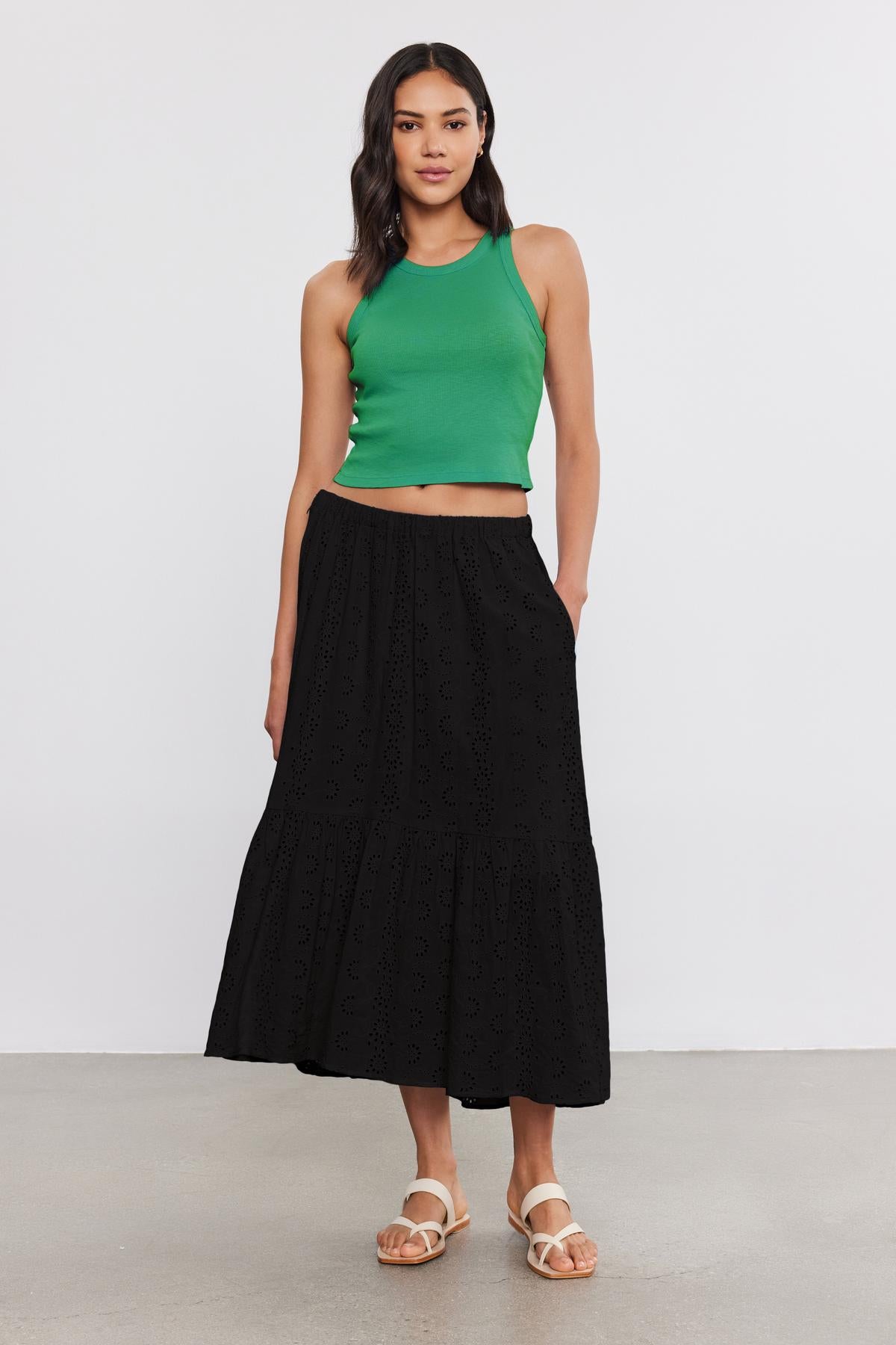   A woman stands against a plain background wearing the HARLA RIBBED CROPPED TANK TOP by Velvet by Graham & Spencer in green, a long black skirt with eyelet details, and white sandals. 