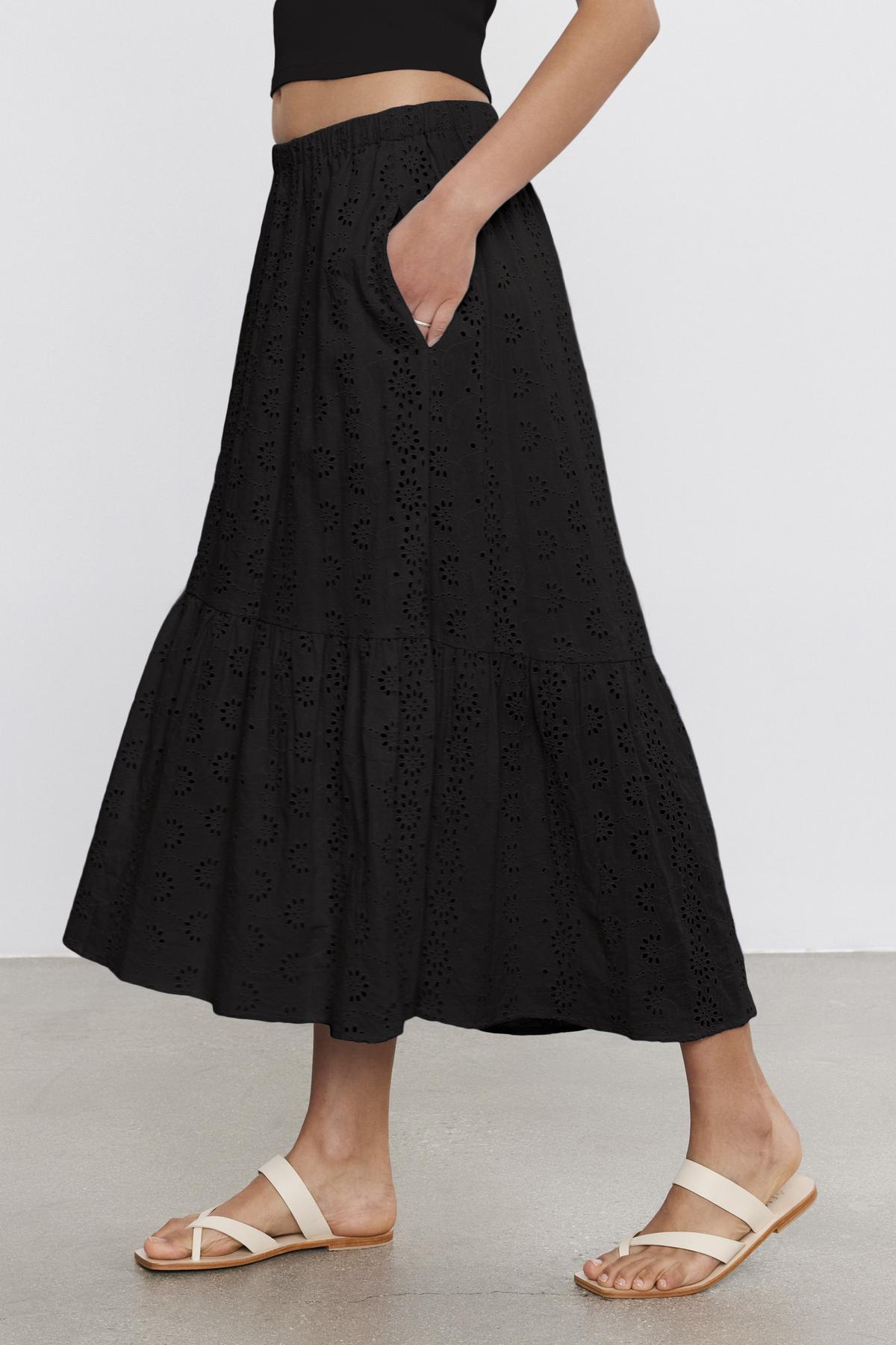 A person wearing the AMELIA SKIRT by Velvet by Graham & Spencer, a black, ankle-length cotton skirt with embroidered details and an elastic waist, paired with a black top and beige sandals stands against a white background.-37934926430401