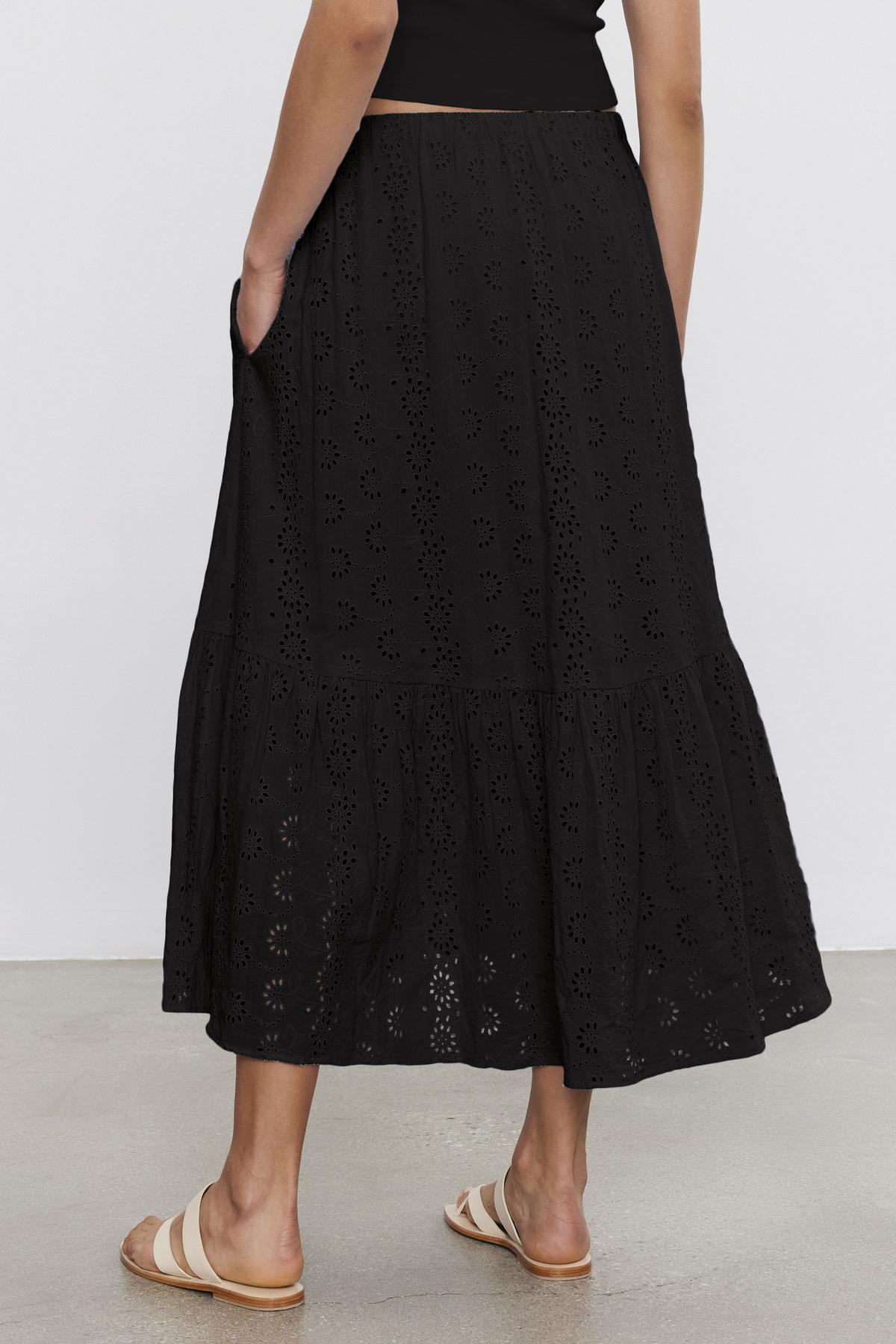   A person wearing the AMELIA SKIRT by Velvet by Graham & Spencer, which is a black, ankle-length broderie anglaise cotton skirt with a tiered design and pockets. The skirt features an elastic waist for comfort. The person is also wearing a black top and beige sandals. 