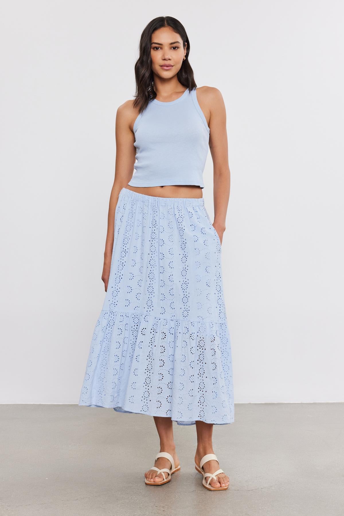 A woman in a light blue tank top and an Amelia skirt by Velvet by Graham & Spencer with a white floral pattern stands confidently in a simple studio setting.-36918838755521