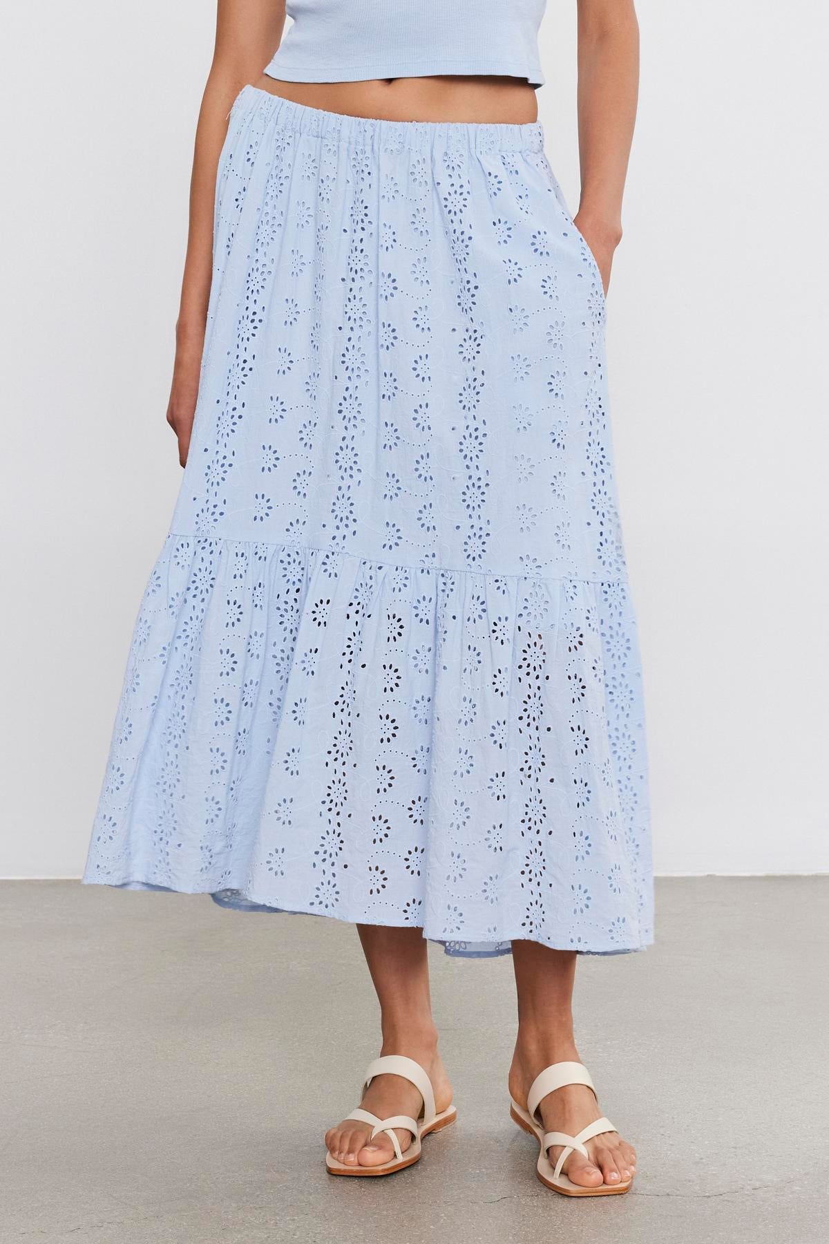   A woman from the waist down wearing a light blue cotton Amelia skirt by Velvet by Graham & Spencer with eyelet details, paired with white sandals, standing against a plain background. 