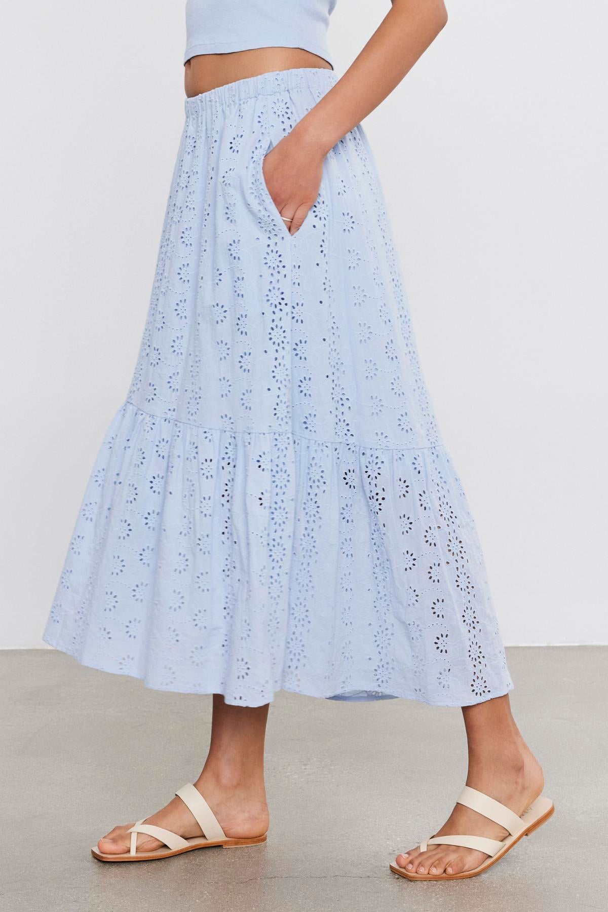   A woman standing wearing a light blue, tiered, broderie anglaise cotton AMELIA SKIRT by Velvet by Graham & Spencer paired with white sandals, cropped view showing from midriff to feet. 