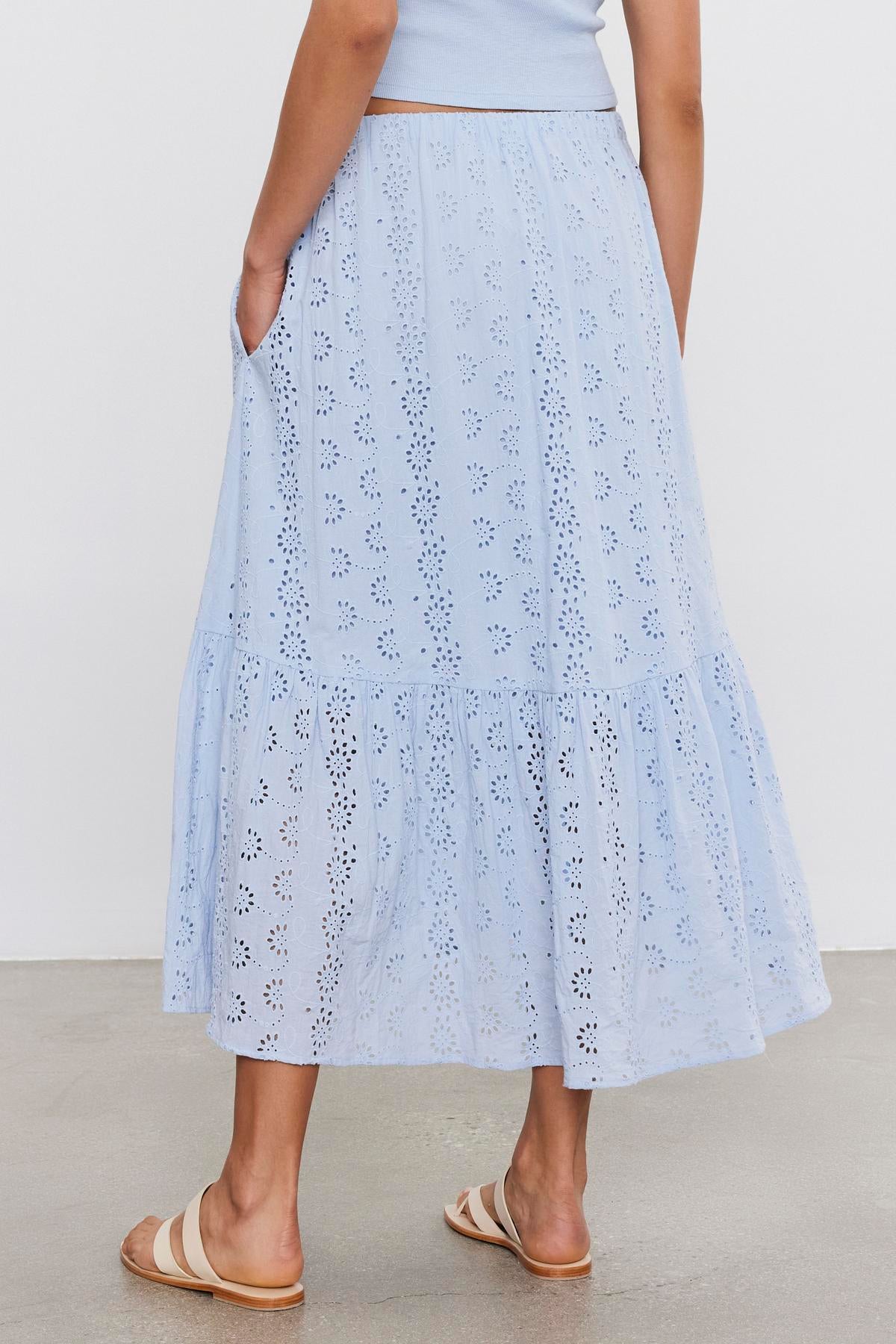   A woman stands showing the back of a light blue, floral eyelet patterned Amelia skirt with a high-low hemline, paired with beige sandals by Velvet by Graham & Spencer. 