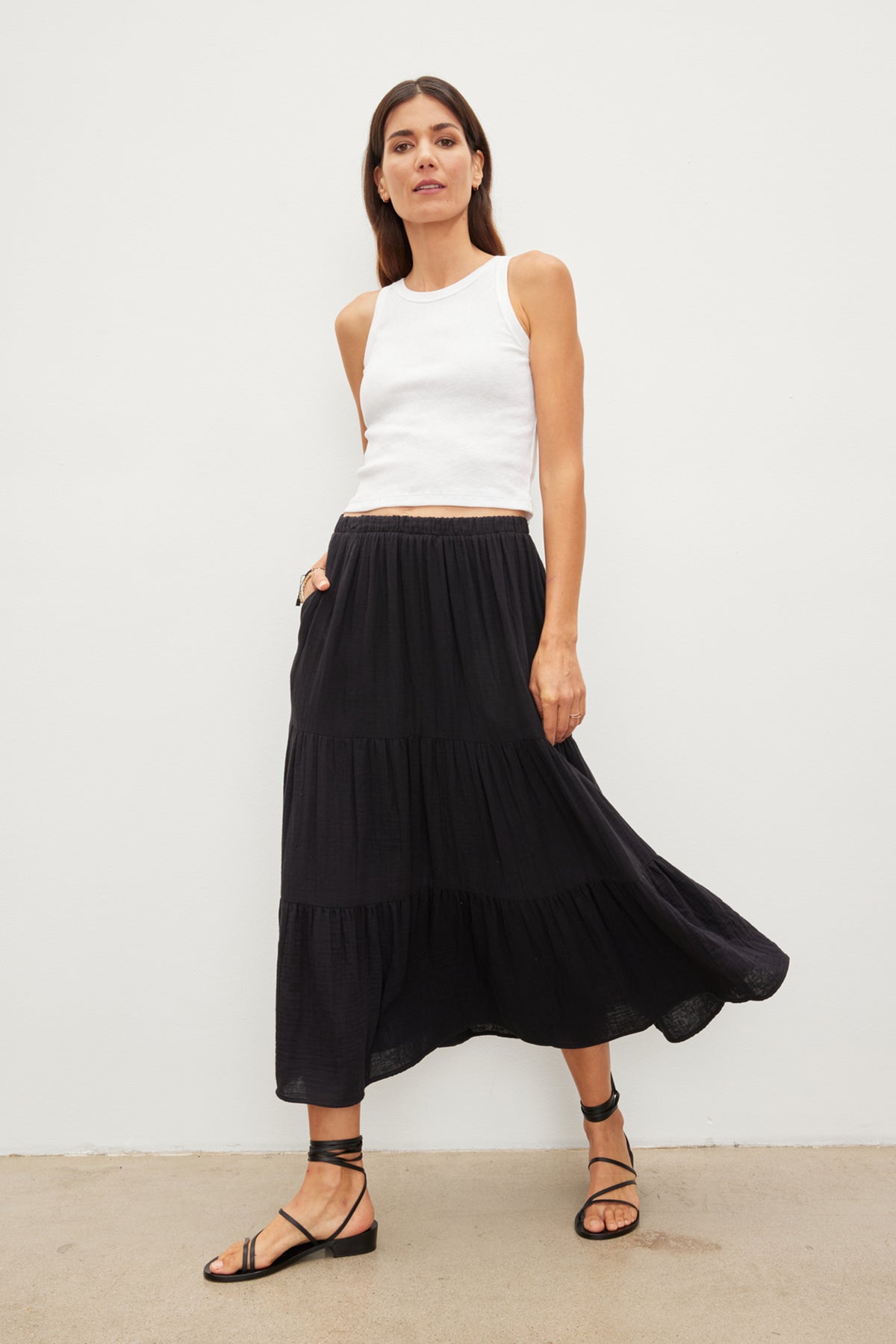 A woman stands against a plain background wearing a white tank top, a DANIELLE COTTON GAUZE TIERED SKIRT by Velvet by Graham & Spencer with an elastic waist, and black strappy sandals.-36443592687809
