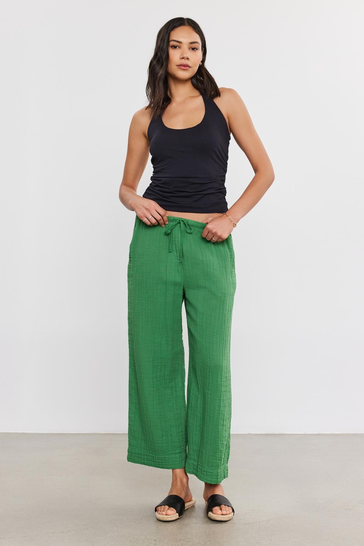   A woman stands confidently, wearing black tank top and Velvet by Graham & Spencer FRANNY COTTON GAUZE PANTS with relaxed legs and slash pockets, paired with black sandals. She's posing in a plain studio setting. 