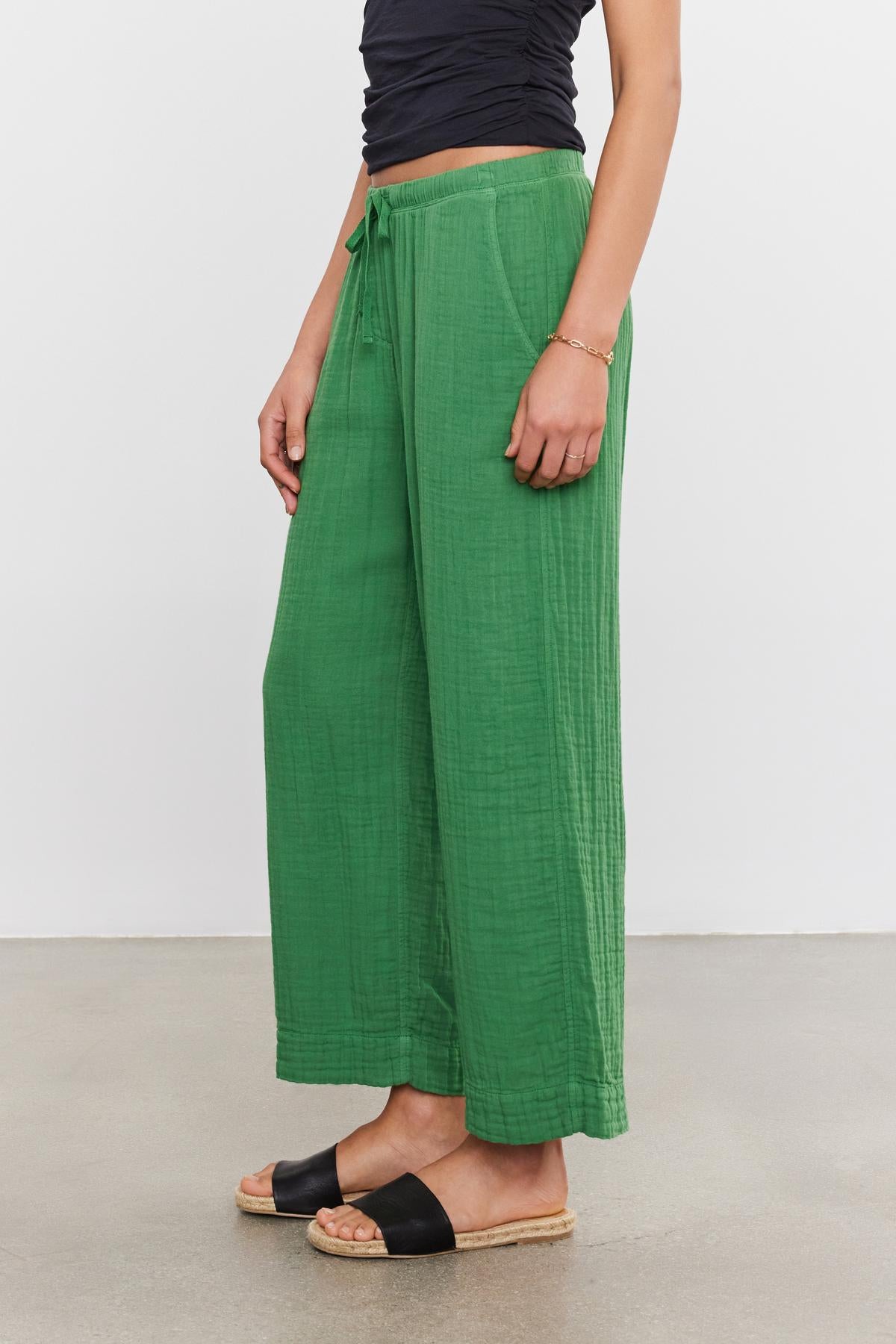 A person wearing green FRANNY COTTON GAUZE PANTS by Velvet by Graham & Spencer with relaxed legs and slash pockets, paired with black slide sandals, standing against a white background.-36917078327489