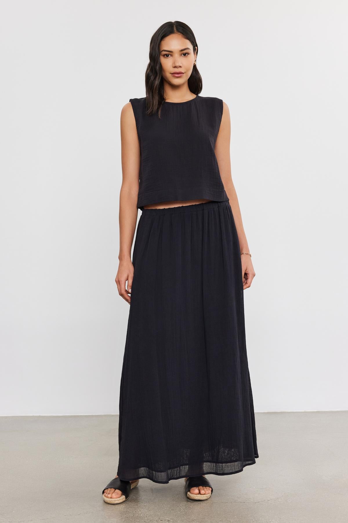A woman in a black sleeveless crop top and a long, flowing Velvet by Graham & Spencer INDY COTTON GAUZE SKIRT stands against a plain background, looking directly at the camera.-36910122533057