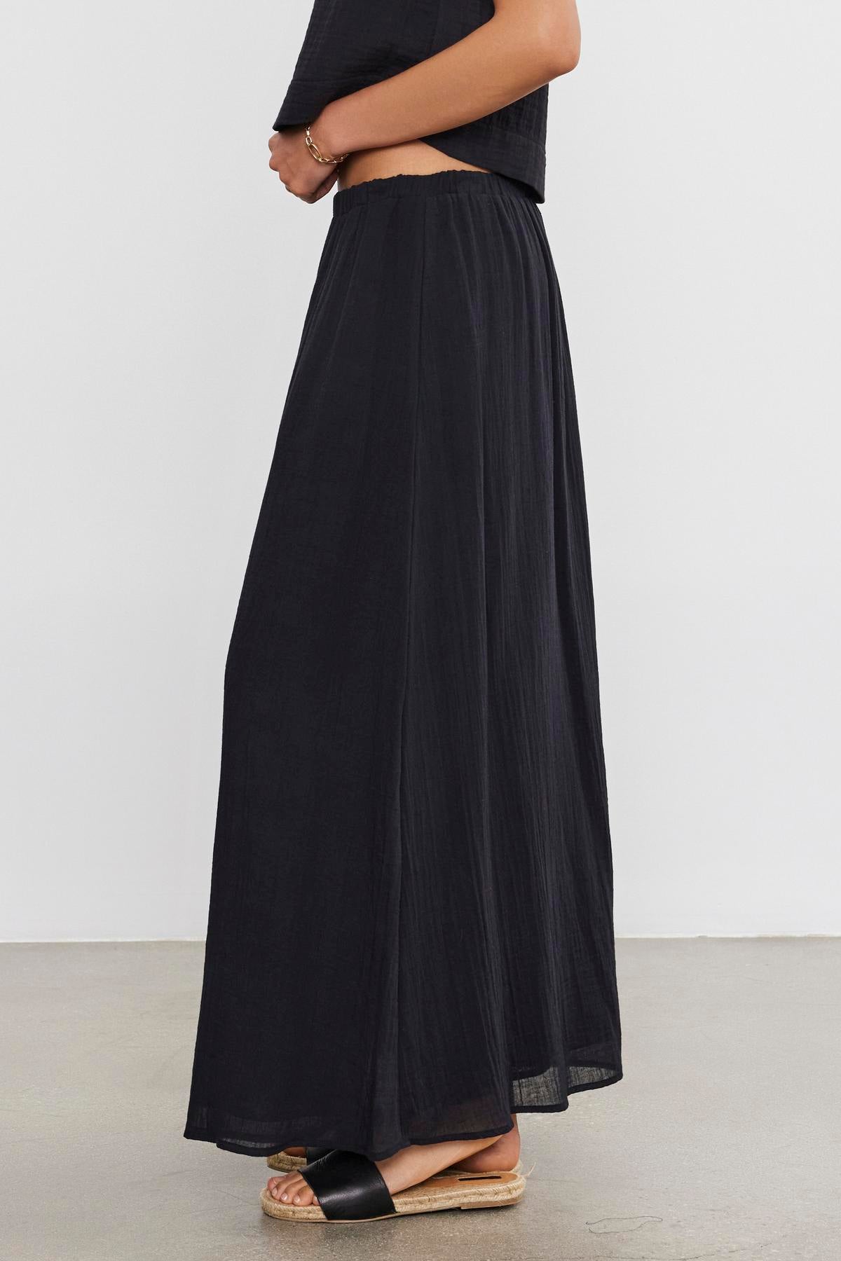 A woman standing in profile wearing a black crop top and a long, flowing INDY COTTON GAUZE SKIRT with sandals by Velvet by Graham & Spencer.-36910123188417