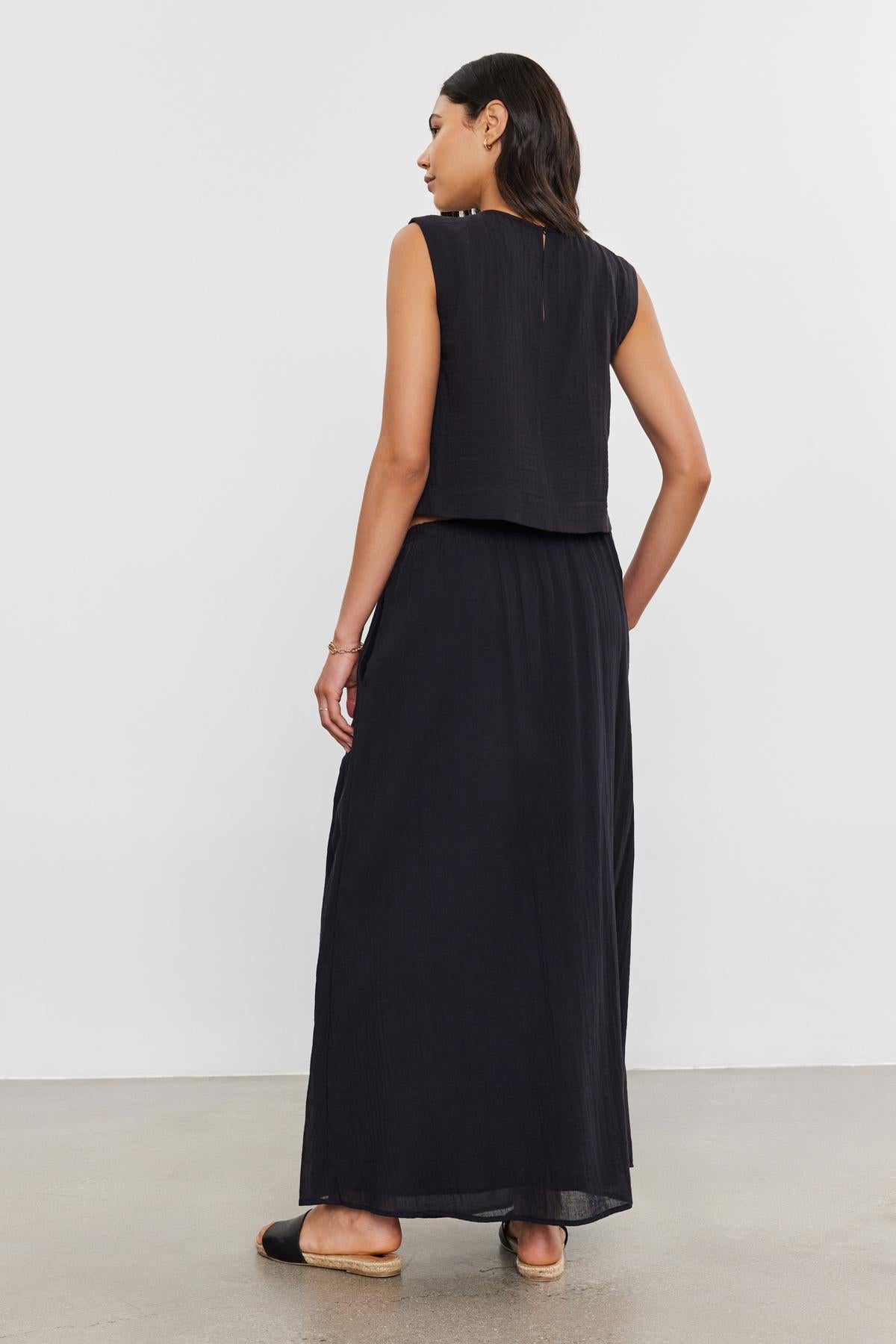 A woman in a black sleeveless maxi dress with an elastic waist skirt, viewed from behind, stands in a plain room, showcasing the back detail of the Velvet by Graham & Spencer INDY COTTON GAUZE SKIRT.-36910123614401