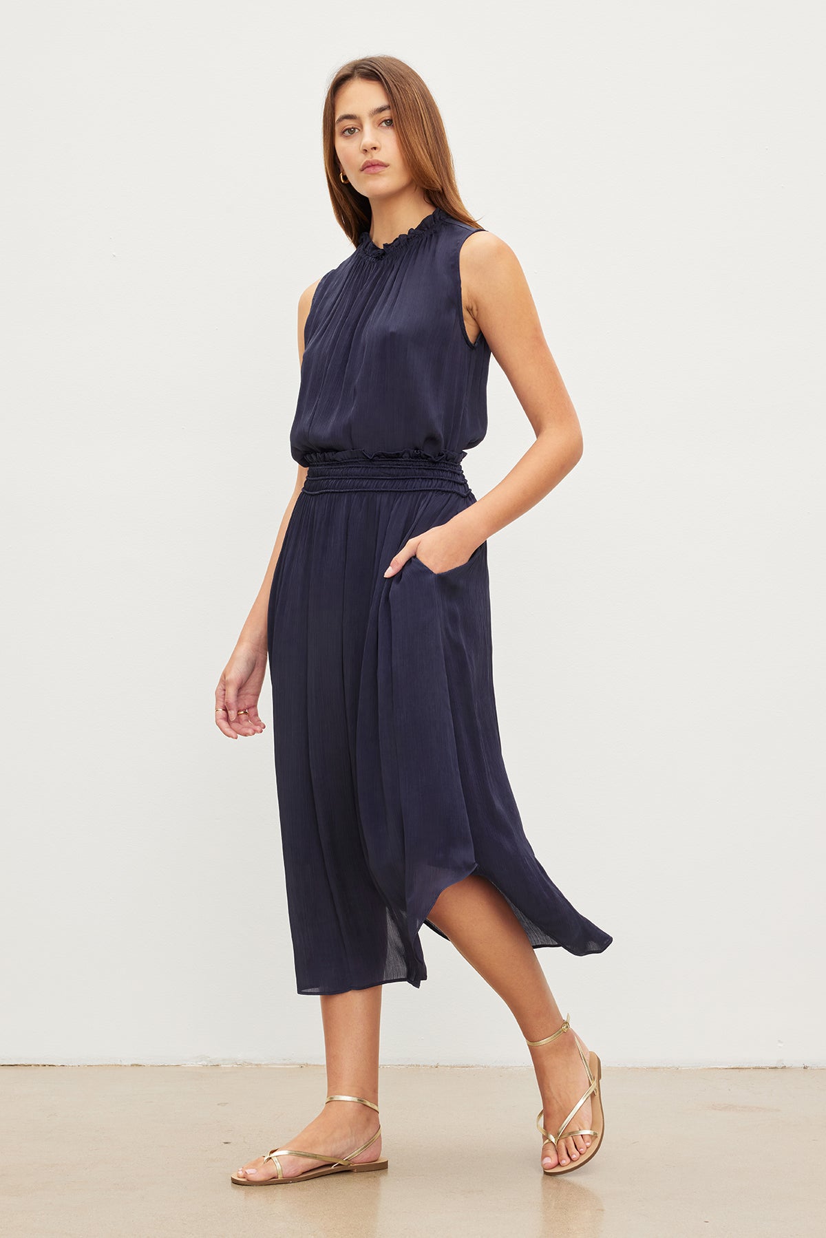 A woman in a navy blue sleeveless dress with an elastic Velvet by Graham & Spencer DIMI smocked skirt waist and gold sandals stands against a plain white background.-36409580028097