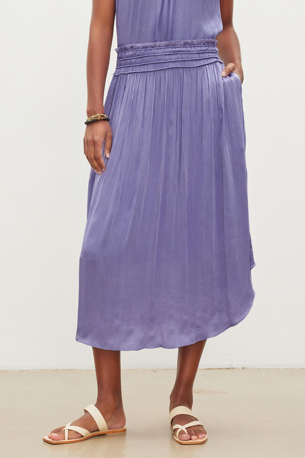   A person wearing a long, purple, crinkled Velvet by Graham & Spencer DIMI SKIRT with one hand in the inseam pocket and beige sandals. The background is white. 