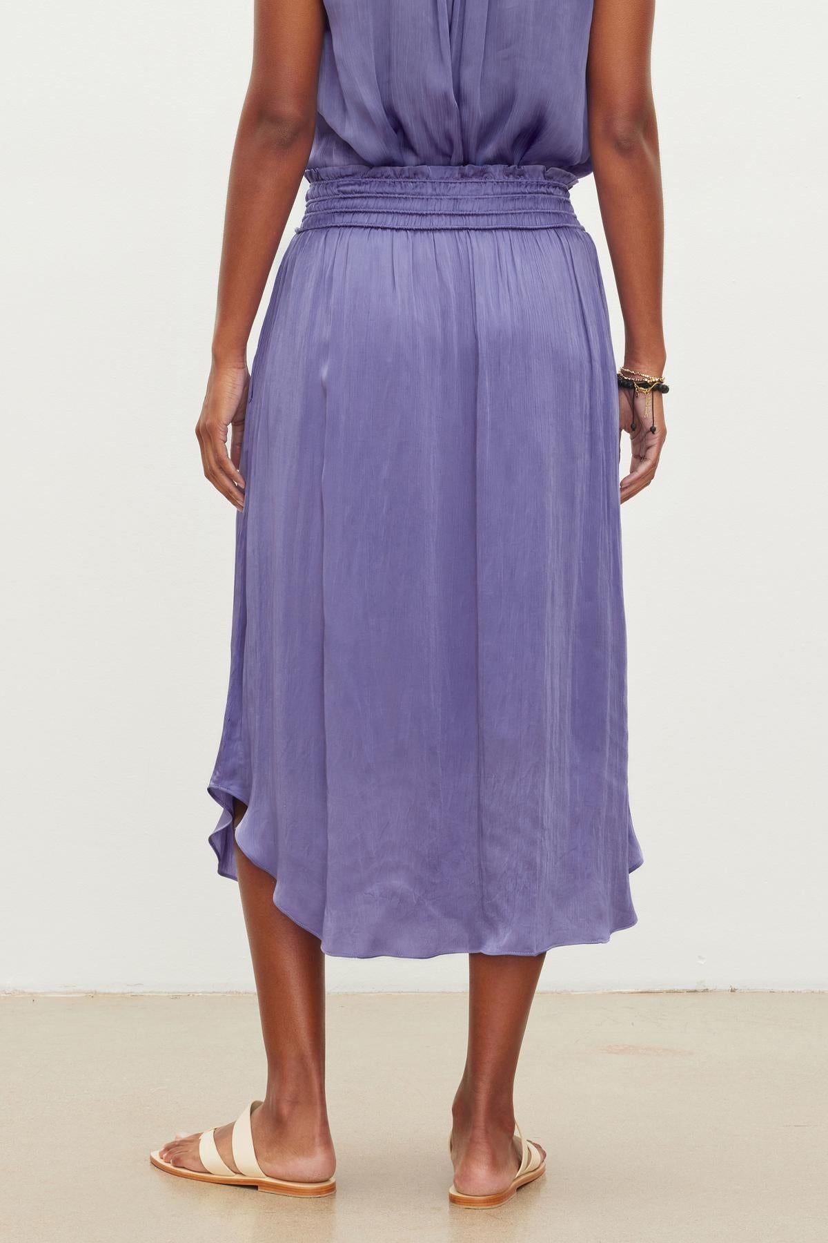   Person standing, wearing a DIMI SKIRT by Velvet by Graham & Spencer made from crinkled viscose with gathered waist and asymmetrical hemline, and tan flat sandals, viewed from the back. 