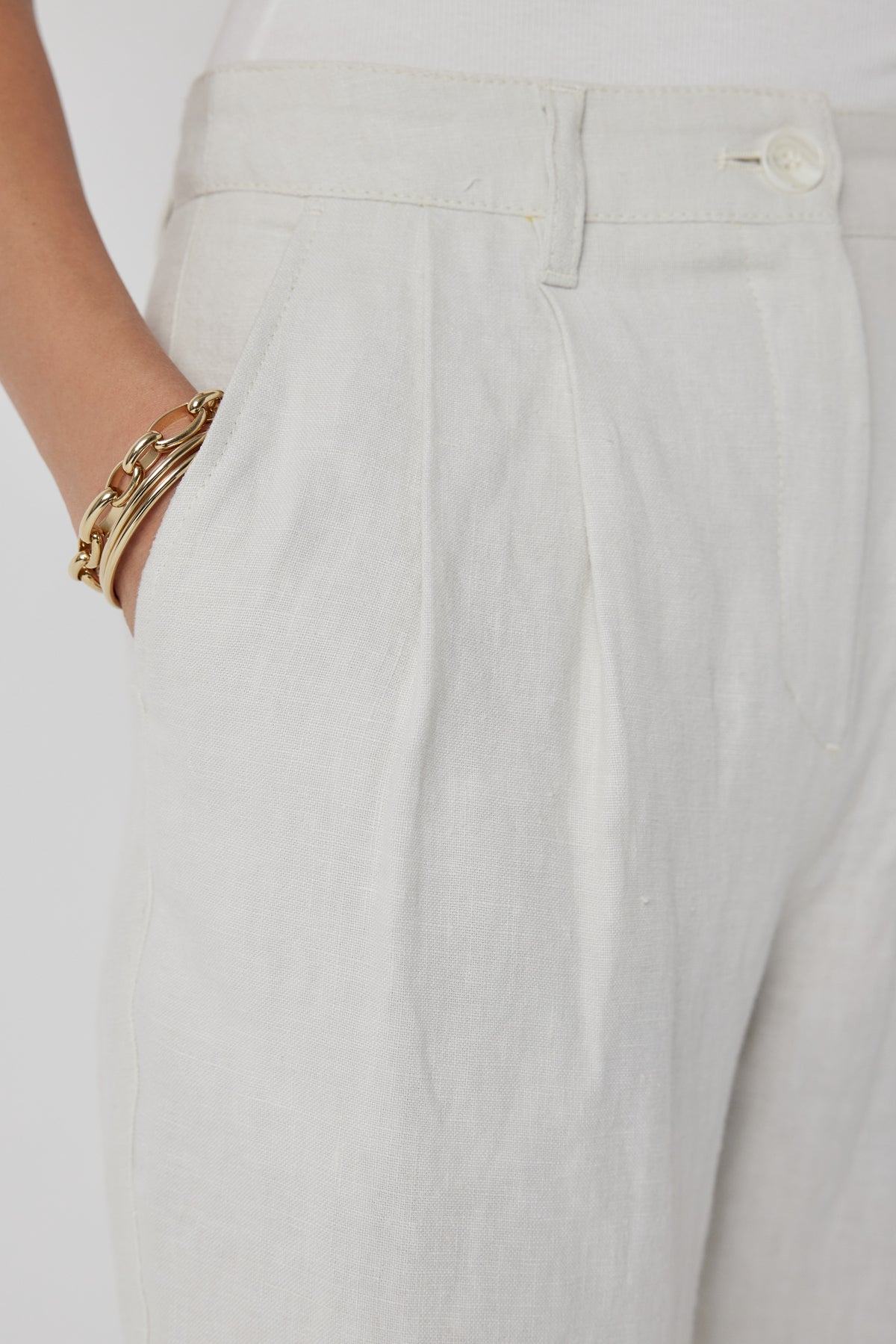   A close-up of a person wearing Velvet by Jenny Graham's POMONA PANT, white heavy-weight linen, straight-leg pants with their hand in the pocket, accessorized with gold bangle bracelets. 