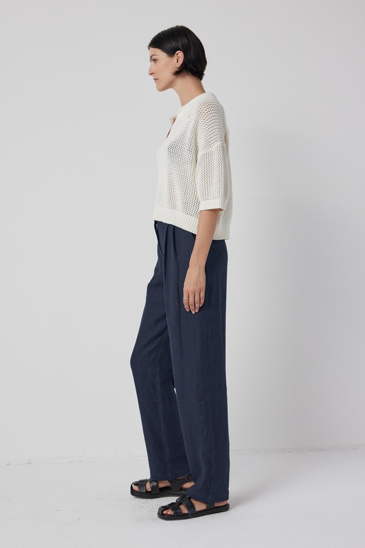 A person with short dark hair stands sideways wearing a white YORK SWEATER POLO by Velvet by Jenny Graham that exudes casual charm, dark blue wide-leg pants, and black sandals.-36463605350593