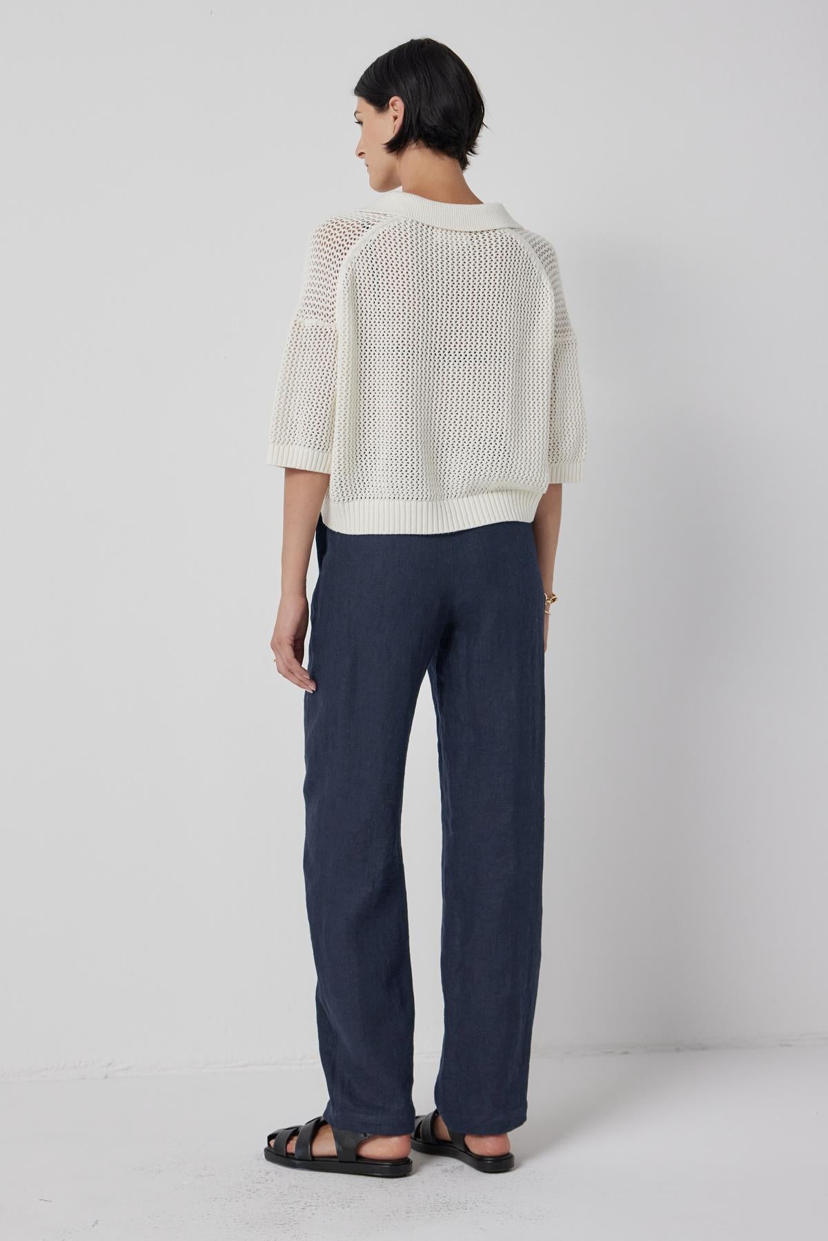 A person with short hair, wearing a York Sweater Polo by Velvet by Jenny Graham, blue pants, and black sandals, stands facing a white wall.-36463605547201