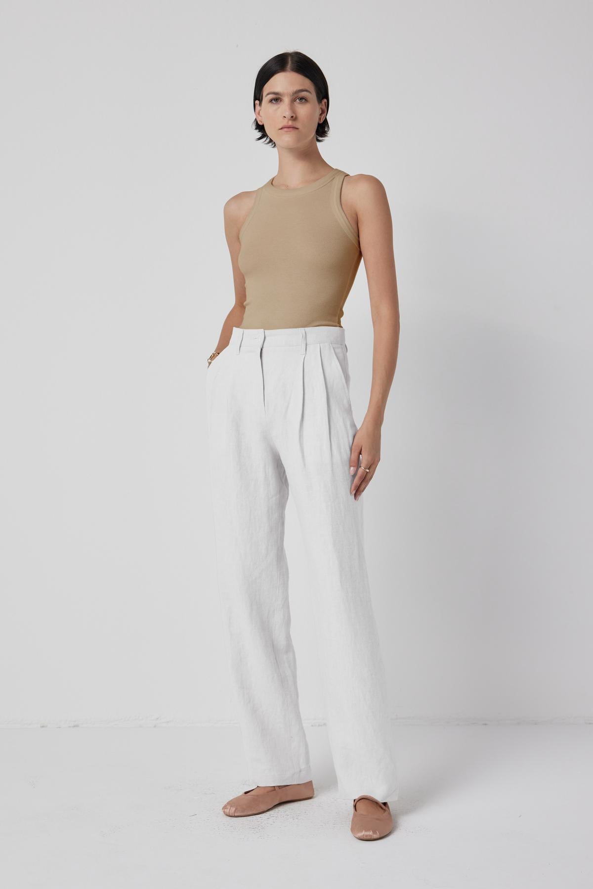   A woman standing against a white background wearing a sleeveless Cruz Tank Top, white trousers, and chic beige flat shoes by Velvet by Jenny Graham. 