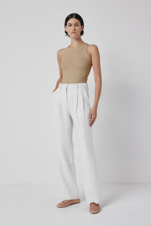 A woman standing against a white background wearing a sleeveless Cruz Tank Top, white trousers, and chic beige flat shoes by Velvet by Jenny Graham.