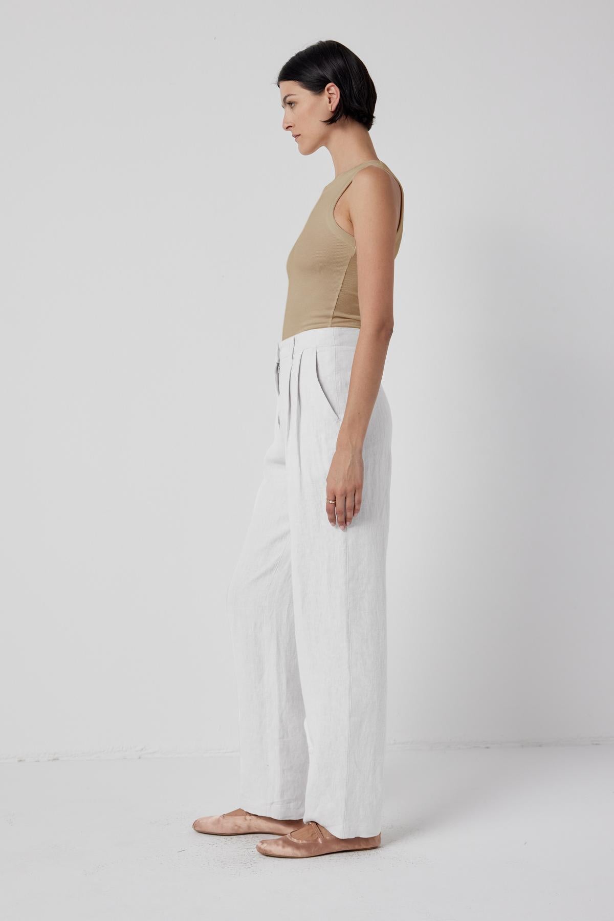   Woman standing in profile wearing a chic beige Velvet by Jenny Graham Cruz tank top and white stretch fit trousers against a white background. 