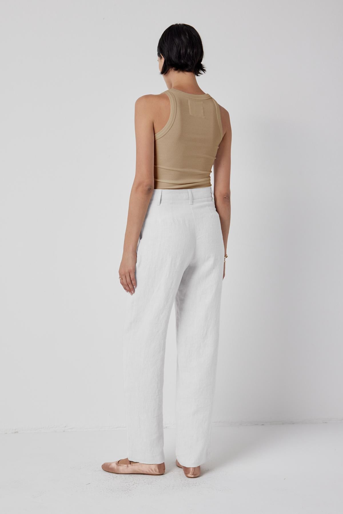   Woman standing in a white studio wearing white pants and a chic beige heavy rib CRUZ tank top by Velvet by Jenny Graham, viewed from behind. 