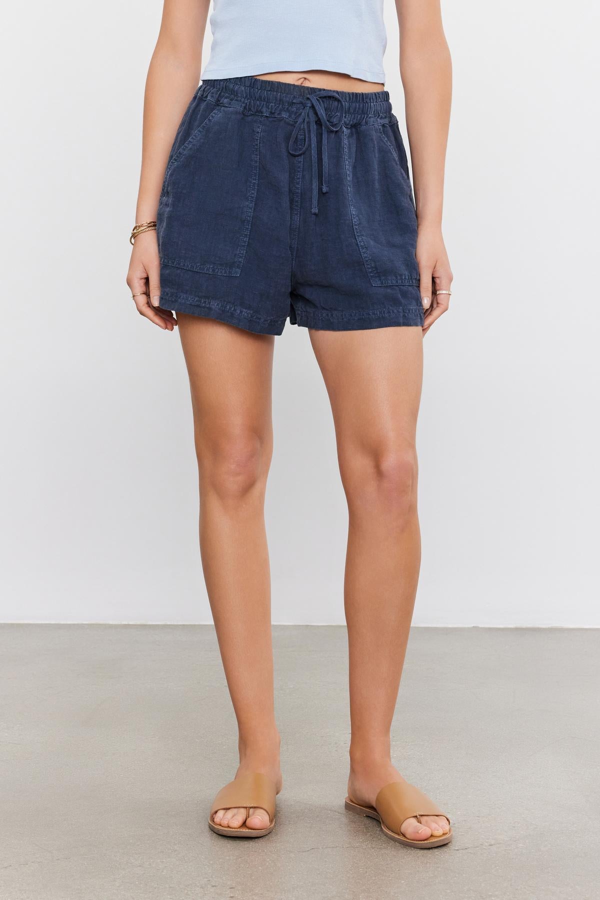   A person wearing a pair of Velvet by Graham & Spencer navy blue drawstring shorts and tan slide sandals, standing against a plain white background. 