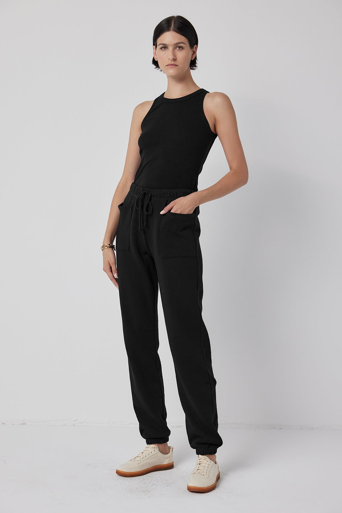   The individual is sporting a black sleeveless top paired with the WESTLAKE SWEATPANT from Velvet by Jenny Graham. They stand against a plain white background, showcasing an outfit that blends sleek style with comfort, crafted from organic cotton fleece and featuring black joggers with an elastic drawstring waist. 