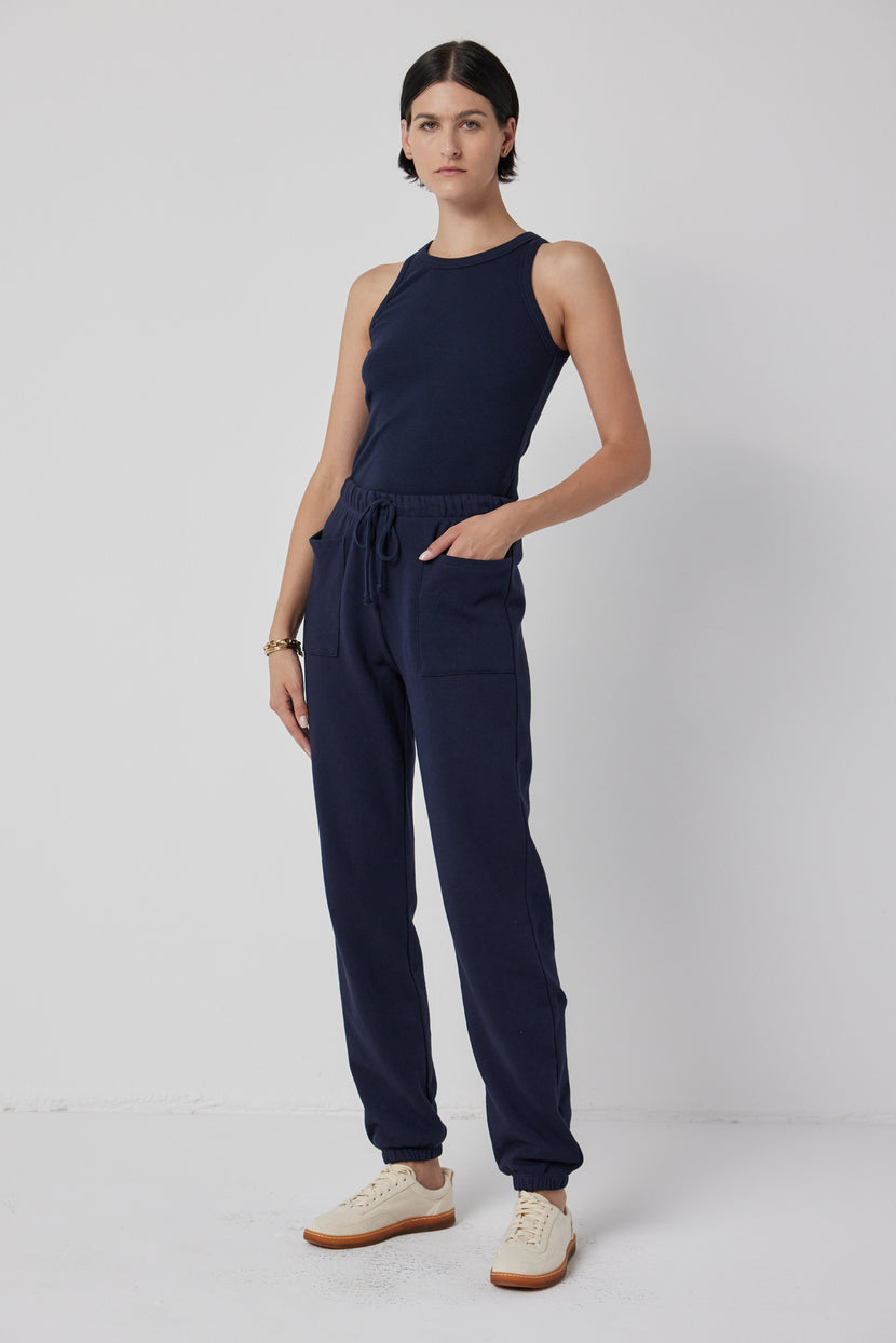 A person stands against a plain white background, dressed in a navy blue sleeveless top and Velvet by Jenny Graham's WESTLAKE SWEATPANT, featuring an elastic drawstring waist and crafted from organic cotton fleece.