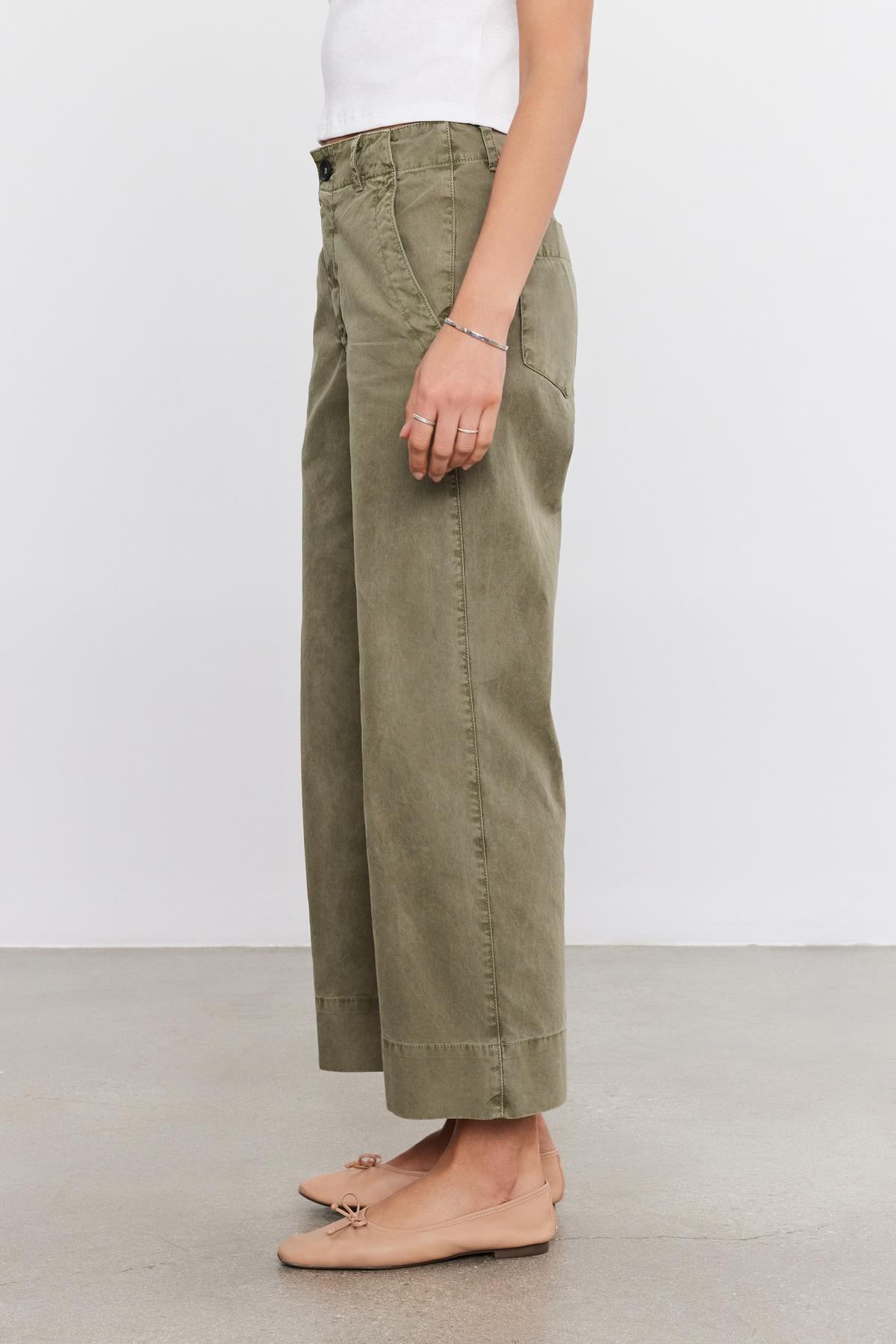   A person wearing a white top, Velvet by Graham & Spencer's green SABINE COTTON CANVAS PANT, and light brown ballet flats stands against a plain background. 