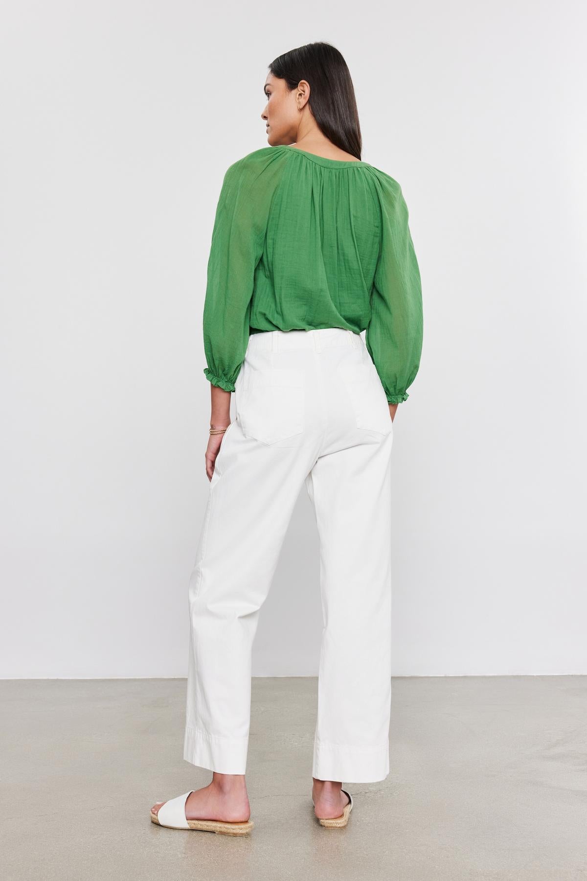   A woman is standing with her back toward the camera, wearing a green long-sleeve blouse, SABINE COTTON CANVAS PANTS by Velvet by Graham & Spencer, and white sandals. 