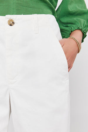 Close-up of a person wearing a green long-sleeve top and SABINE COTTON CANVAS PANT by Velvet by Graham & Spencer, their right hand in the pant pocket. The white pants have a versatile fit with a wide-leg design and buttoned closure.