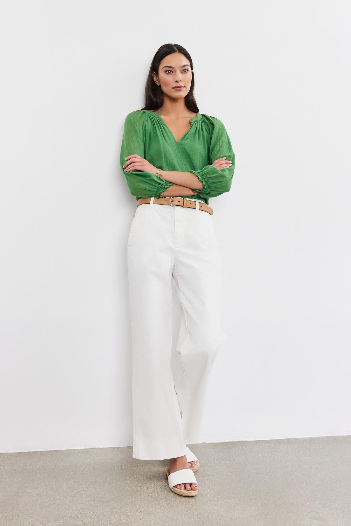   A woman stands against a plain white wall, wearing a green blouse, the SABINE Cotton Canvas Pant by Velvet by Graham & Spencer with a wide leg design, a tan belt, and white sandals. She has her arms crossed and looks straight ahead. 