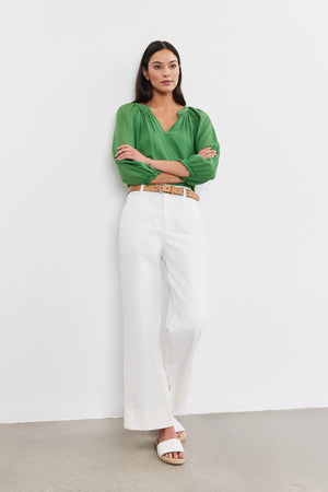 A woman stands against a plain white wall, wearing a green blouse, the SABINE Cotton Canvas Pant by Velvet by Graham & Spencer with a wide leg design, a tan belt, and white sandals. She has her arms crossed and looks straight ahead.