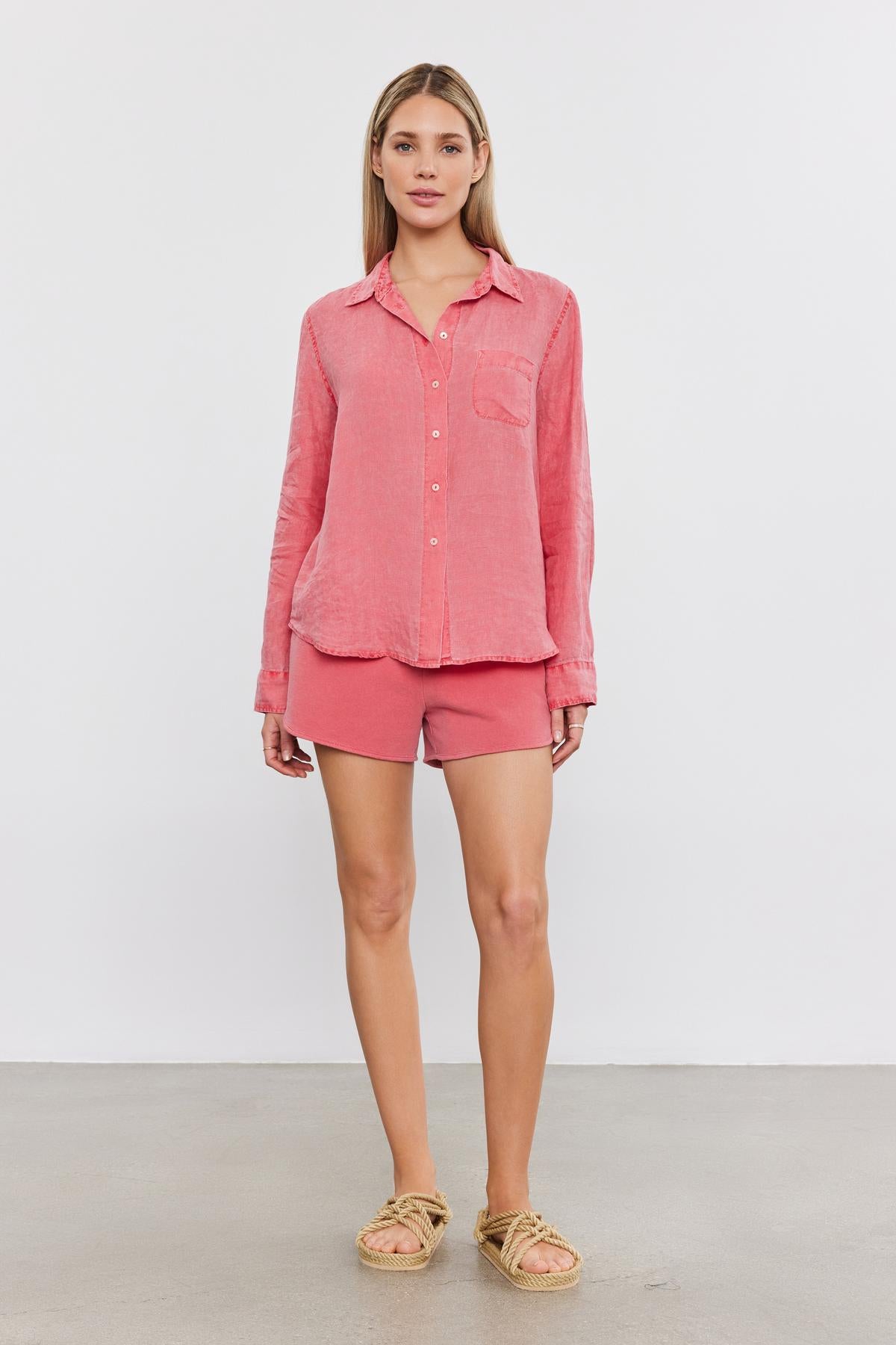   A woman stands facing the camera, wearing a coral pink buttoned shirt, matching Velvet by Graham & Spencer Presely shorts, and beige woven sandals. She has blonde hair and a neutral expression. 