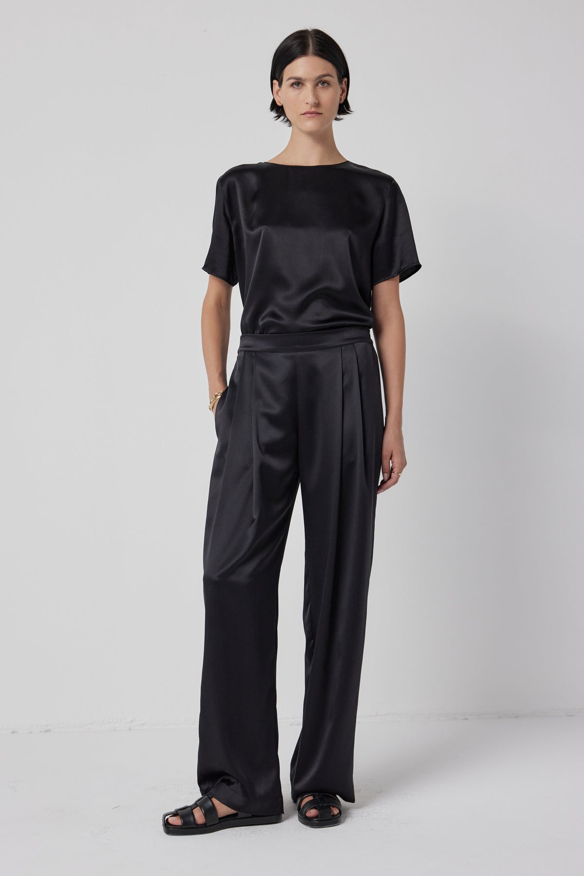 A woman wearing a black silk charmeuse Soho top and MANHATTAN PANT trousers with black sandals against a white background.-36463440593089