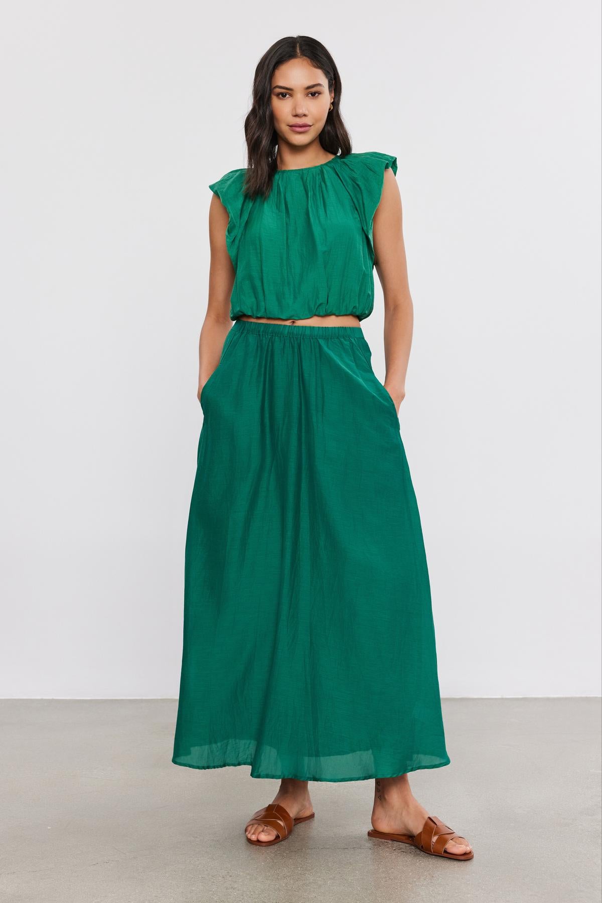 A woman wearing a green sleeveless top and a Velvet by Graham & Spencer MARIELA SKIRT stands on a plain background, paired with brown sandals.-36917062303937