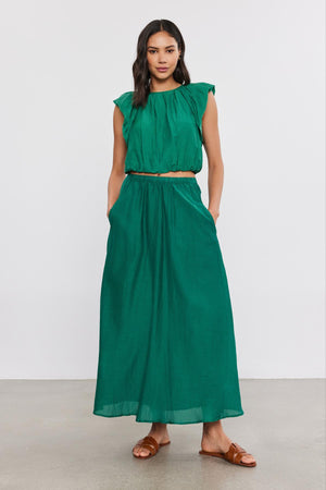 A woman wearing a green sleeveless top and a Velvet by Graham & Spencer MARIELA SKIRT stands on a plain background, paired with brown sandals.