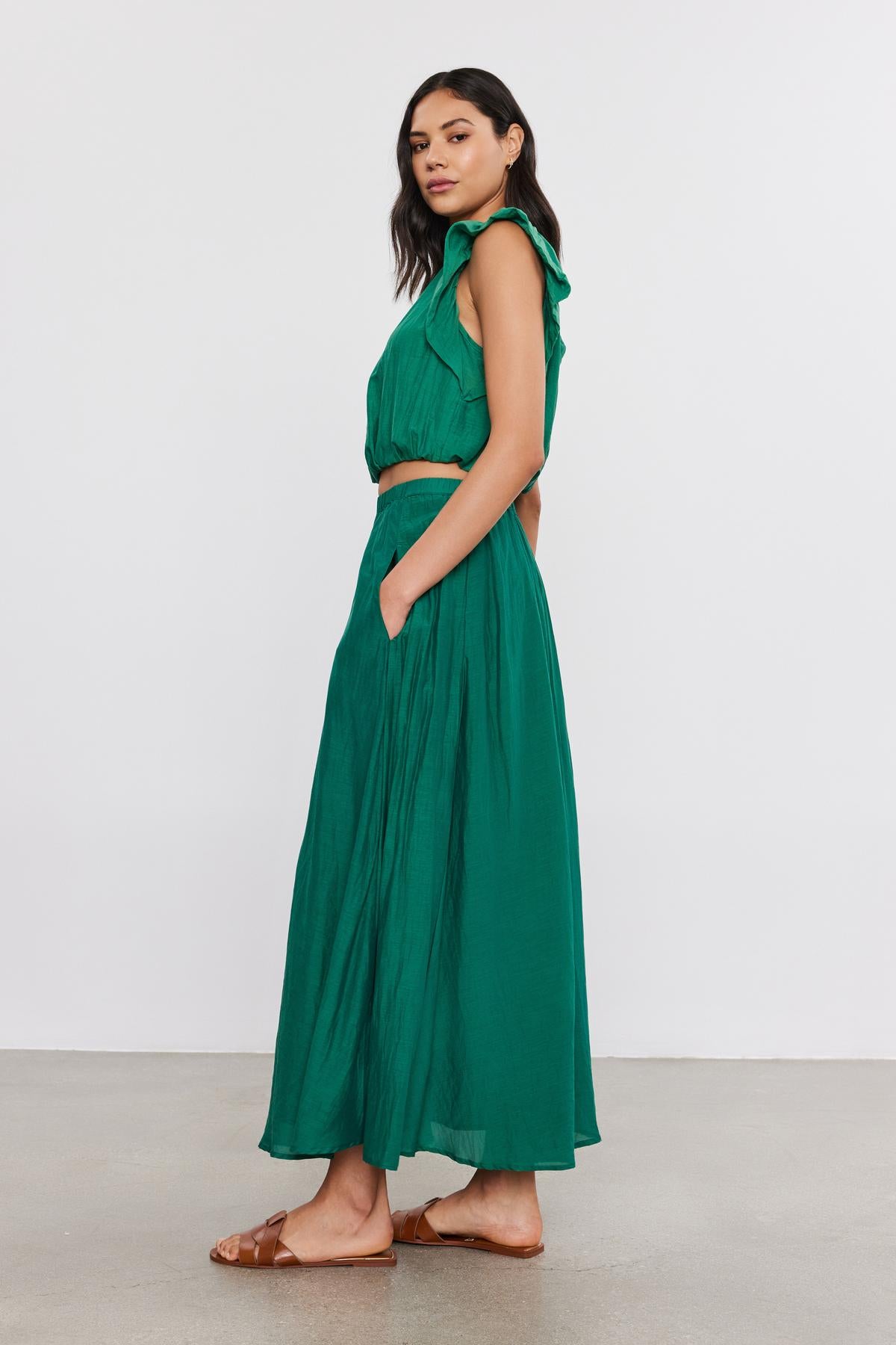   Woman in a green sleeveless lined Mariela skirt and brown sandals, standing sideways, looking over her shoulder against a neutral background. 