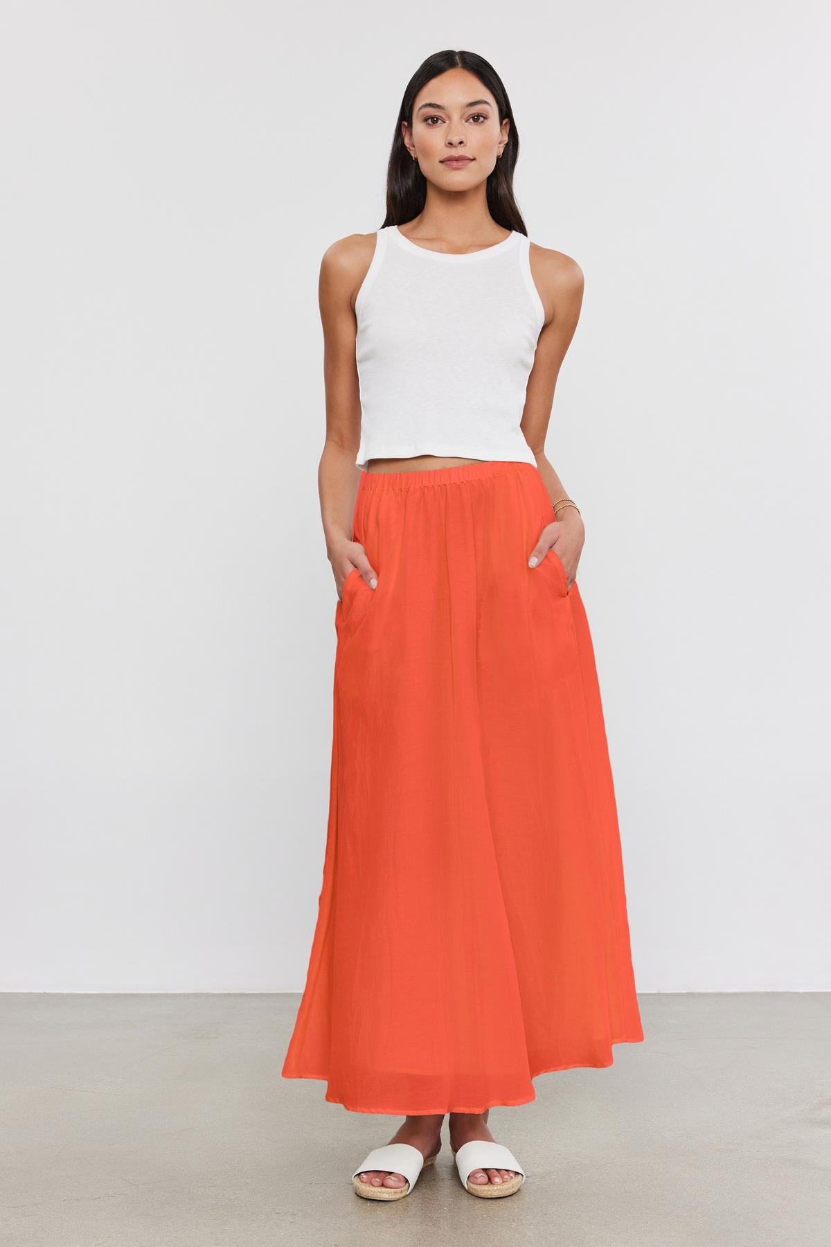   A woman stands against a plain background, wearing a white sleeveless top, the MARIELA SKIRT by Velvet by Graham & Spencer – an orange lined maxi skirt with pockets made from silk cotton voile – and white sandals. 