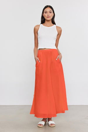 A woman stands against a plain background, wearing a white sleeveless top, the MARIELA SKIRT by Velvet by Graham & Spencer – an orange lined maxi skirt with pockets made from silk cotton voile – and white sandals.