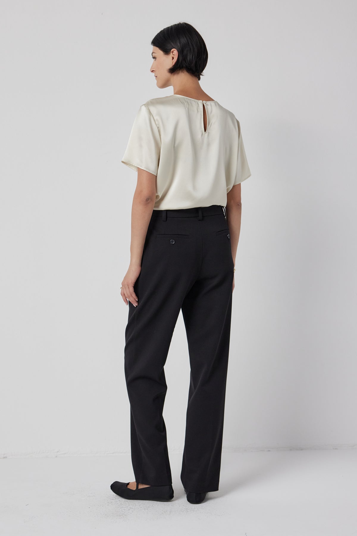 A woman stands facing away from the camera, showcasing the back of a Velvet by Jenny Graham Pasadena top and black trousers, radiating timeless elegance.-36463793995969