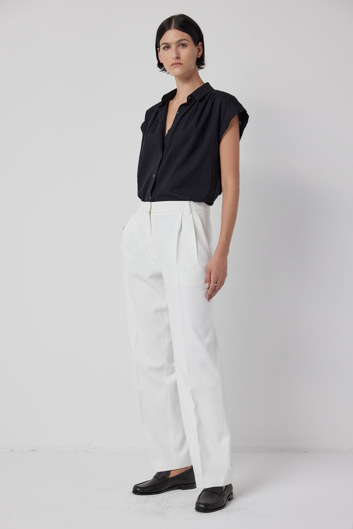   A woman standing against a white backdrop, wearing a black blouse, white Velvet by Jenny Graham relaxed fit BUNDY PANT trousers, and black loafers. 