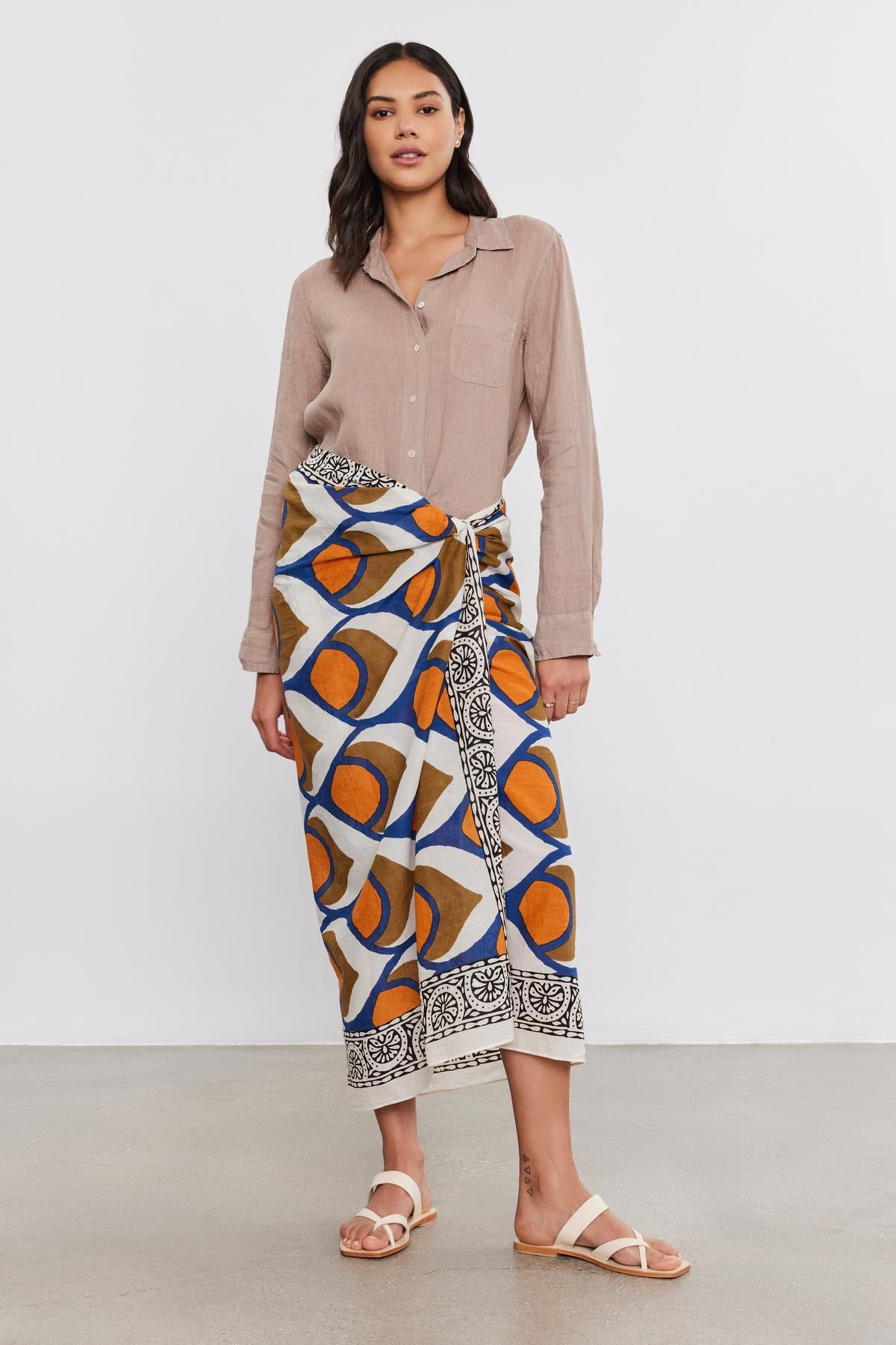   A woman in a studio poses wearing a Velvet by Graham & Spencer NATALIA LINEN BUTTON-UP SHIRT, a colorful patterned skirt, and white sandals. 