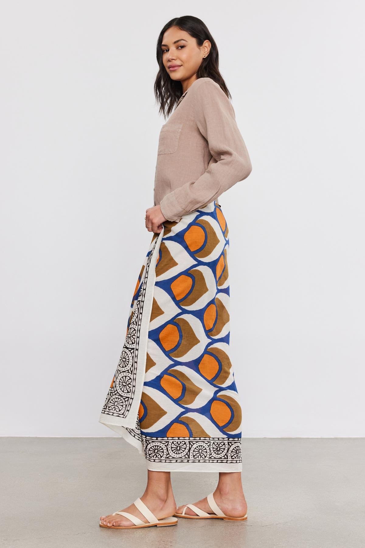 Woman standing sideways, wearing a beige blouse, colorful printed skirt, and white sandals by Velvet by Graham & Spencer, holding a SARONG WRAP bag, looking over her shoulder.-36752949149889