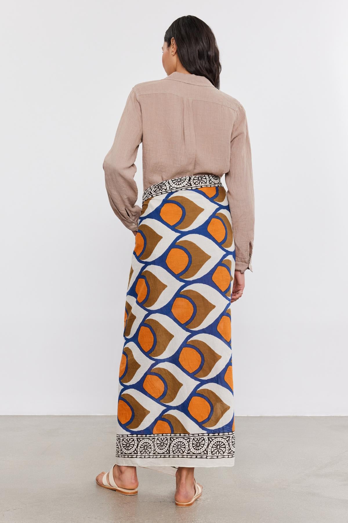 A woman standing with her back to the camera, wearing a beige blouse and a colorful geometric-patterned SARONG WRAP skirt from Velvet by Graham & Spencer, paired with tan sandals.-36752949182657