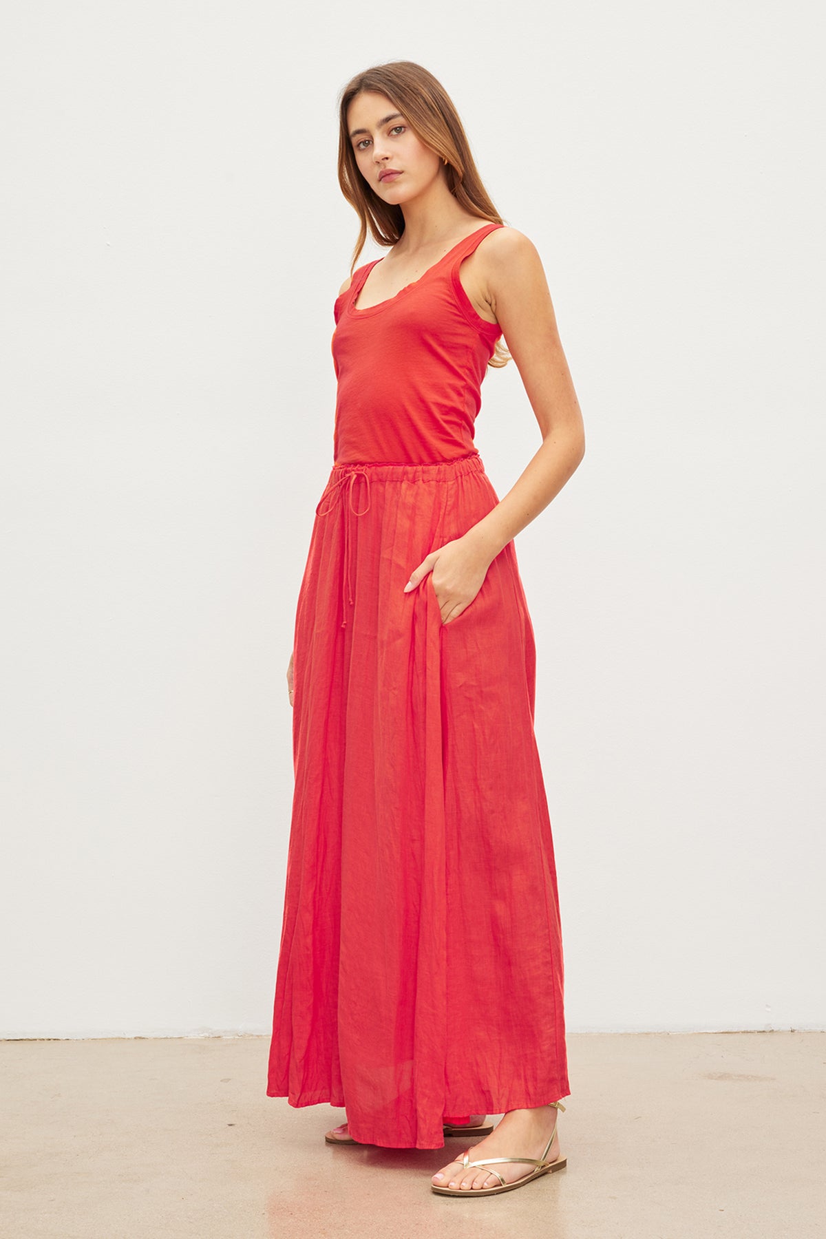   A woman wearing the BAILEY LINEN MAXI SKIRT in red from Velvet by Graham & Spencer and beige sandals poses against a plain white background. She has one hand in her pocket and is looking at the camera. 