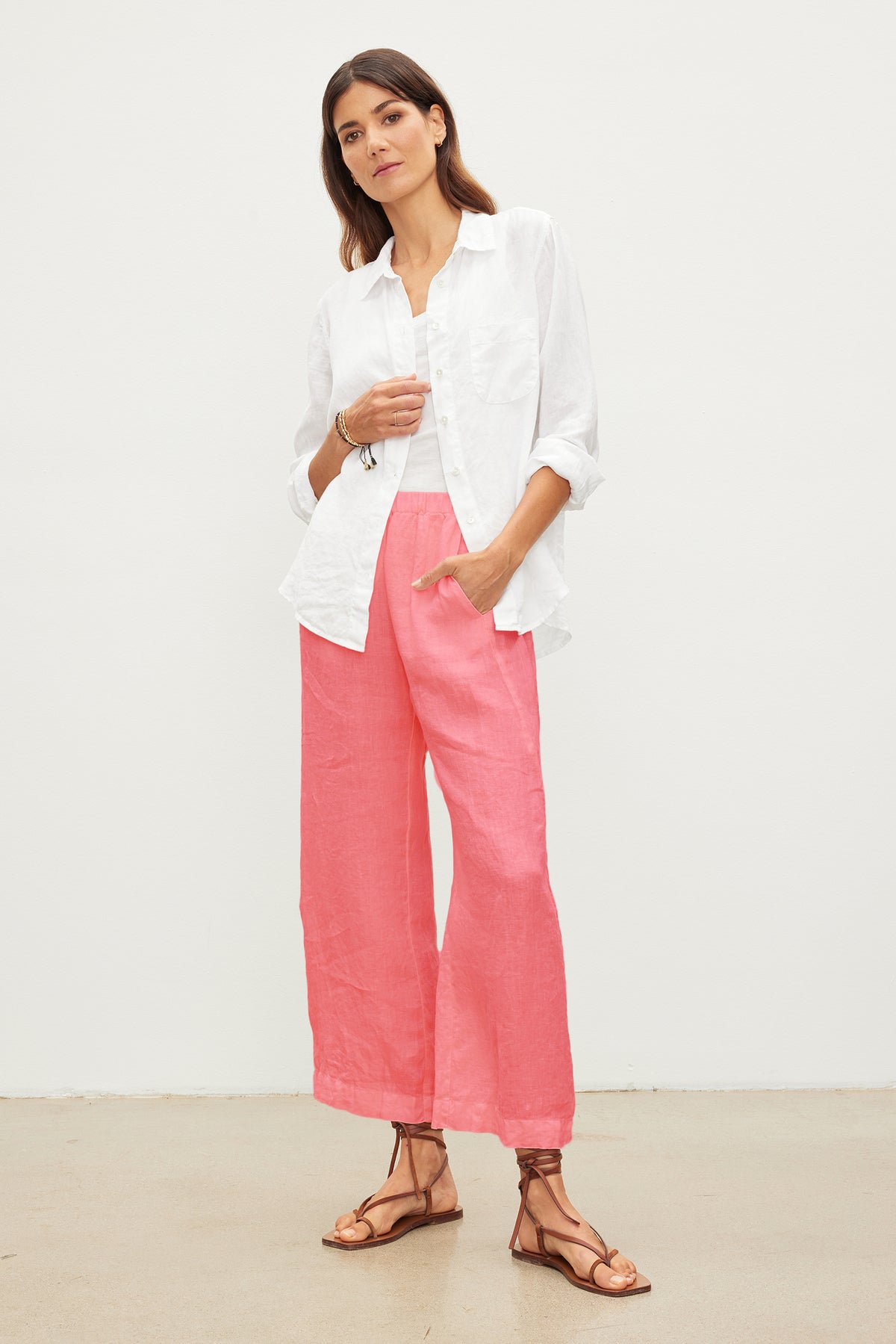 A woman stands against a plain white background, wearing a white button-up shirt and pink Velvet by Graham & Spencer LOLA LINEN PANT with an elastic waist. She has brown hair and is wearing sandals.-37260951126209